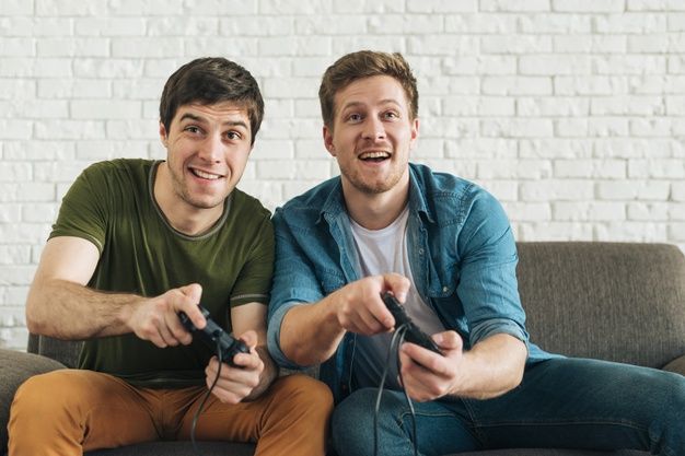 Friends playing video games on couch - Two happy male friends sitting on sofa playing video game with ...