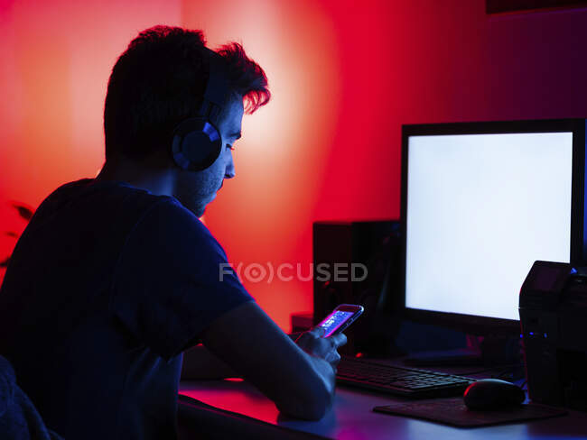 Gamer focused on smartphone screen in dark room - Side view focused young male in casual shirt and wireless ...